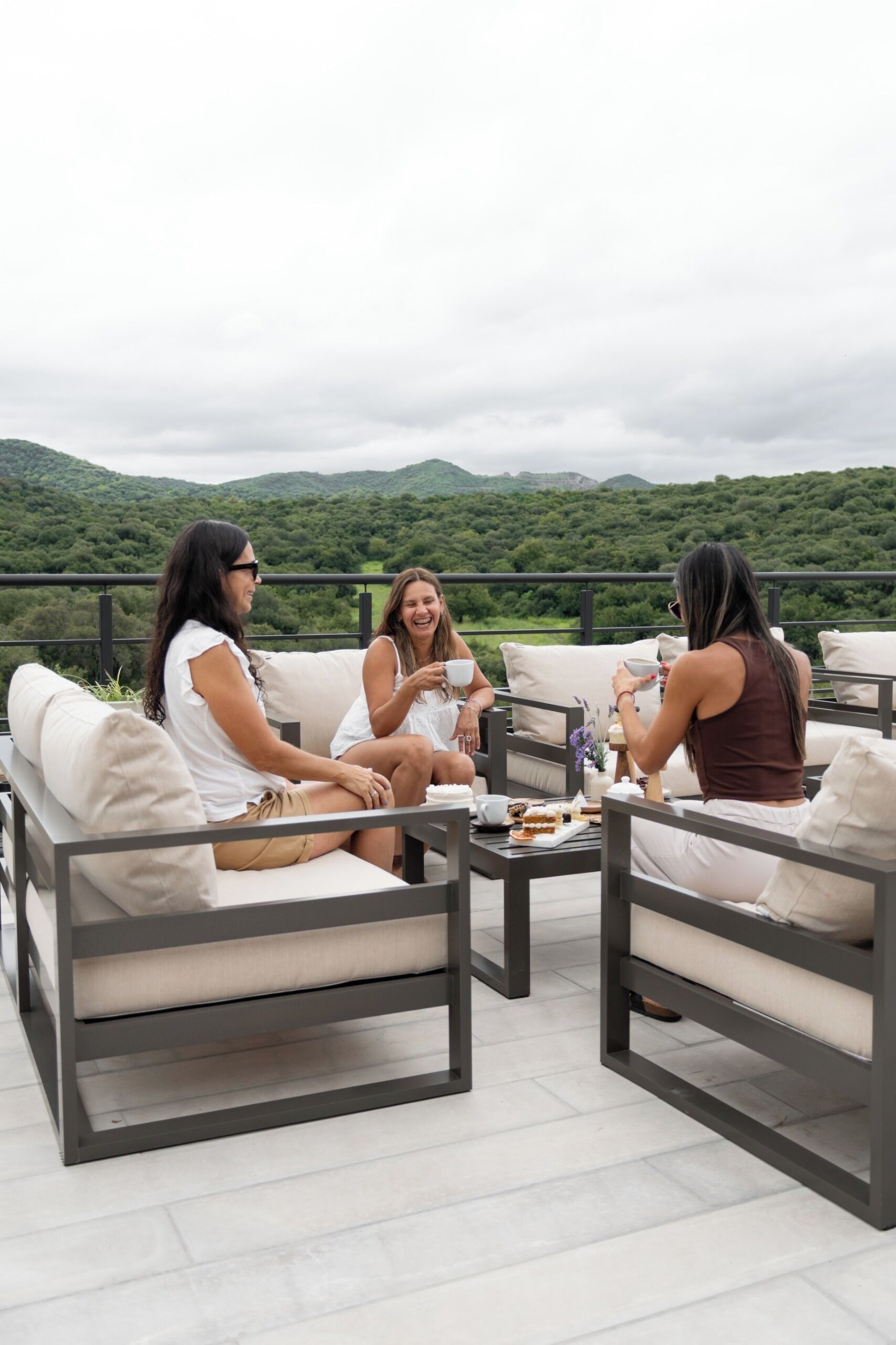 A terrace with three people seated on sofas, a table with food, and a view of green hills.