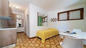A compact room with a bed, a small table, a chair, a kitchenette, and a mirror. - Pal Friul - Villa Marinella (Mola)
