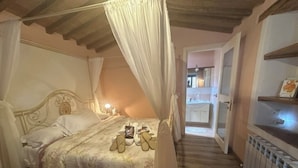 Une chambre avec un lit à baldaquin, une table de chevet et une commode. Il y a un radiateur au sol et une porte qui donne sur une autre pièce.