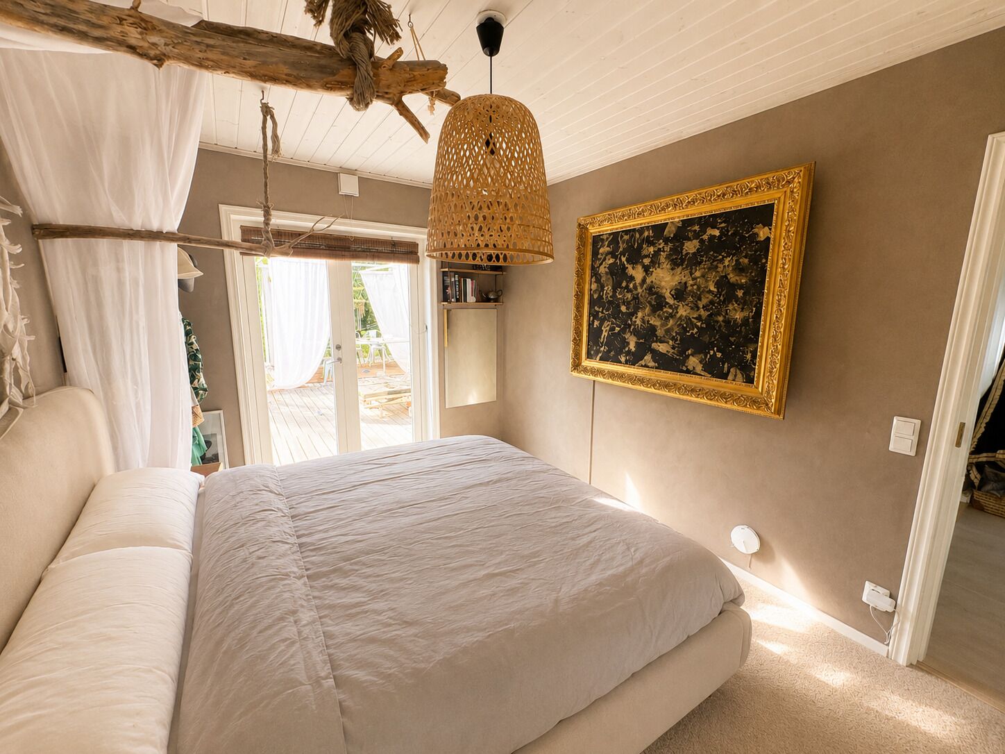 Une chambre avec un grand lit, une lampe en rotin suspendue, un tableau encadré et une vue sur l’extérieur.