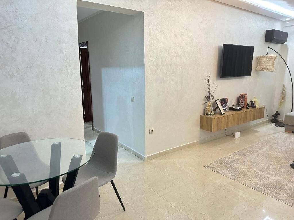 A modern living room with a glass dining table, grey chairs, a mounted TV, and a wooden console with decorative items.