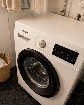 Una lavadora Siemens en un baño con suelo de baldosas, una cesta y un tendedero.
