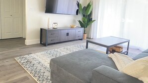 Un soggiorno moderno con un divano grigio, un tavolino da caffè, una televisione a schermo piatto montata a parete, un mobile contenitore grigio e una grande pianta verde.