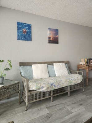 Ein gemütliches Zimmer mit einem Korbssofa, einem kleinen Beistelltisch mit Pflanze und zwei gerahmten Bildern an der Wand.
