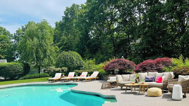 A swimming pool with lounge chairs and a seating area with cushions and tables. - Luxury World Cup Escape–Beautiful home w/Pool & Hot Tub - 9 miles from Gillette! (Mansfield)