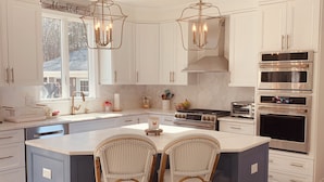 A spacious kitchen with white cabinets, a large island with two wicker chairs, stainless steel appliances, and pendant lighting. - Luxury World Cup Escape–Beautiful home w/Pool & Hot Tub - 9 miles from Gillette! (Mansfield)