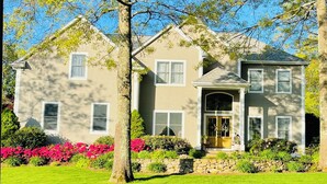 A two-story house with a stone foundation, a front entrance with a door, and a well-maintained lawn with pink flowers. - Luxury World Cup Escape–Beautiful home w/Pool & Hot Tub - 9 miles from Gillette! (Mansfield)