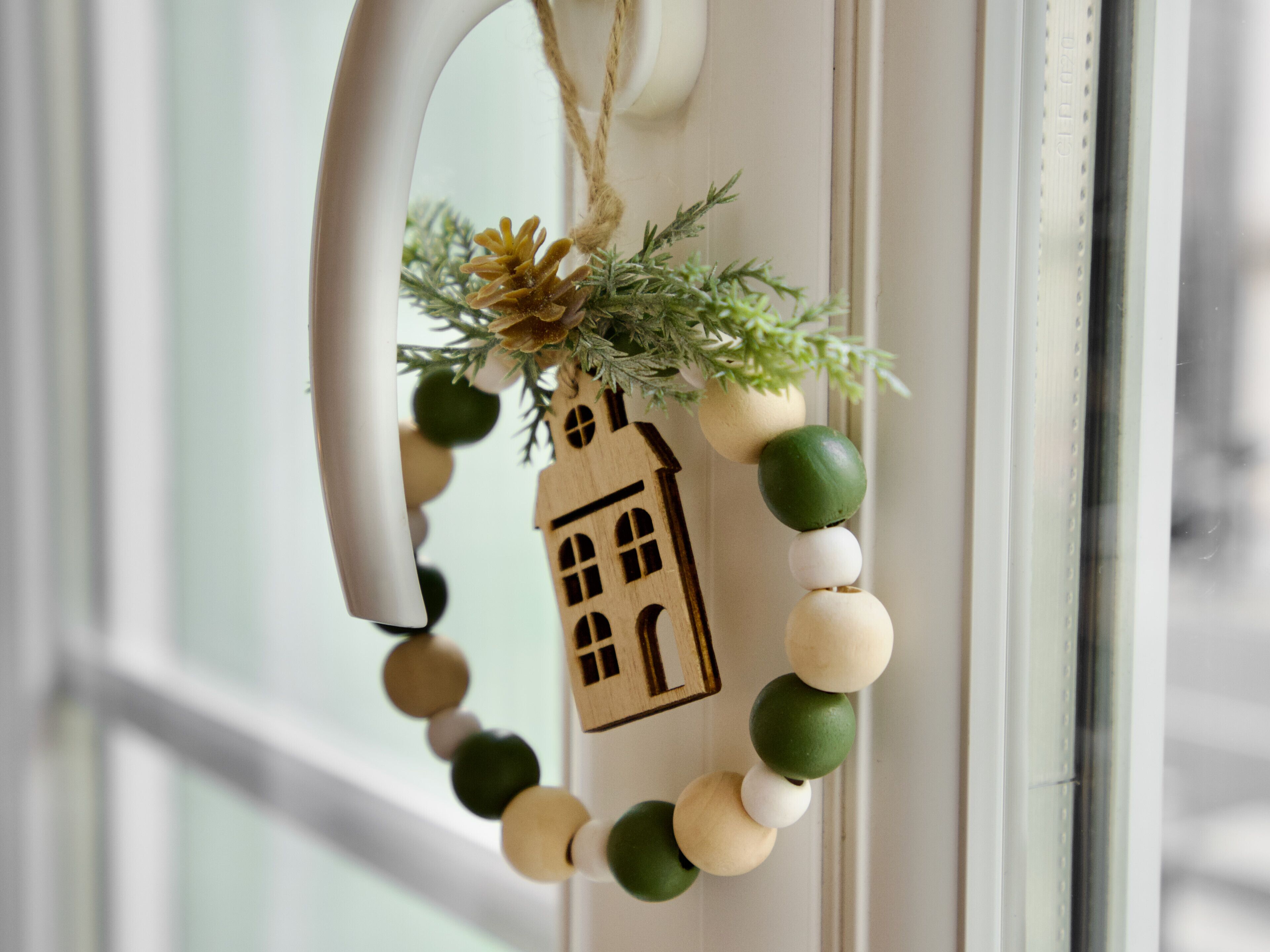 A wooden house ornament with a string of beads and greenery, hanging on a white door.