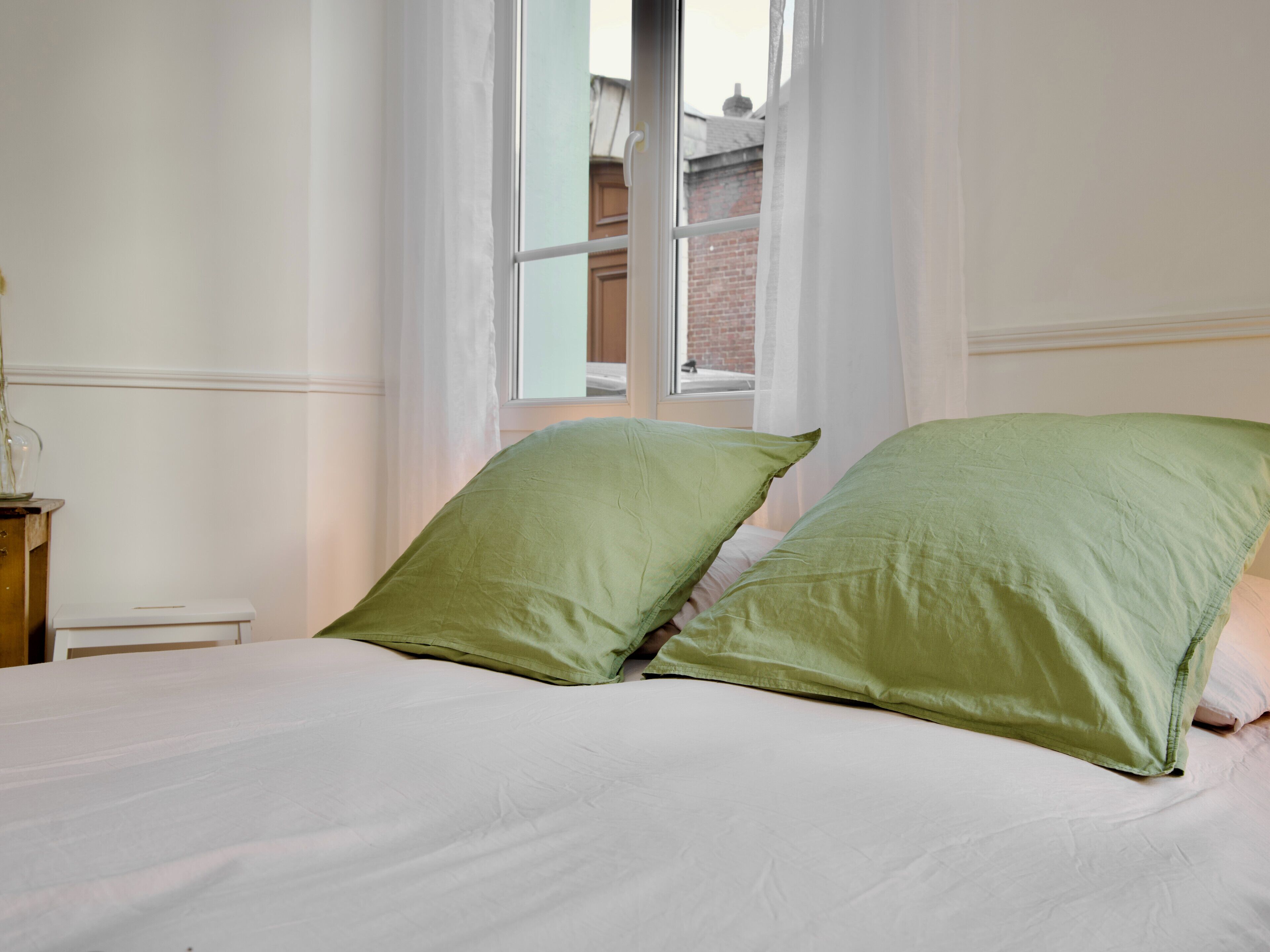 A bed with two green pillows, a wooden nightstand, and a window with a view of a brick building.