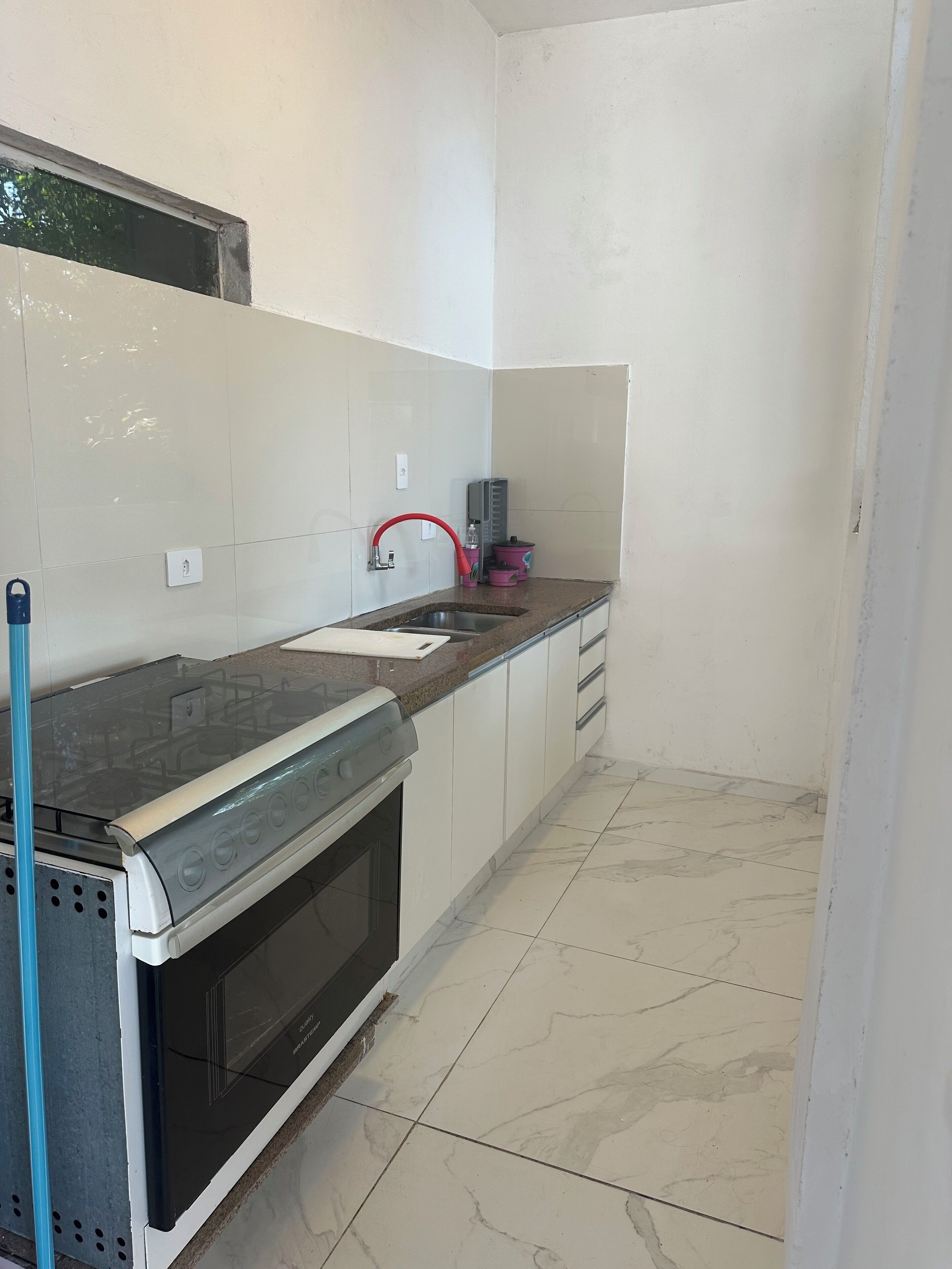 A compact kitchen with white cabinets, a black stove, a sink with a red faucet, and a marble-tiled floor.