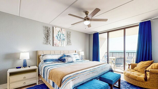 Ein modernes Schlafzimmer mit einem großen Bett, einem Deckenventilator, einem Nachttisch, einem blauen Teppich, einem gelben Sessel und einem Balkon mit blauen Vorhängen.