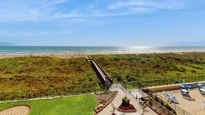 Blick auf den Strand vom Hotelbalkon mit einem Weg zum Strand, einer umzäunten Terrasse mit Liegestühlen und einer Rasenfläche mit einem kleinen Garten.