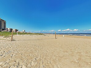 Ein Strand mit Volleyballnetz, sandigem Untergrund und Gebäuden im Hintergrund.