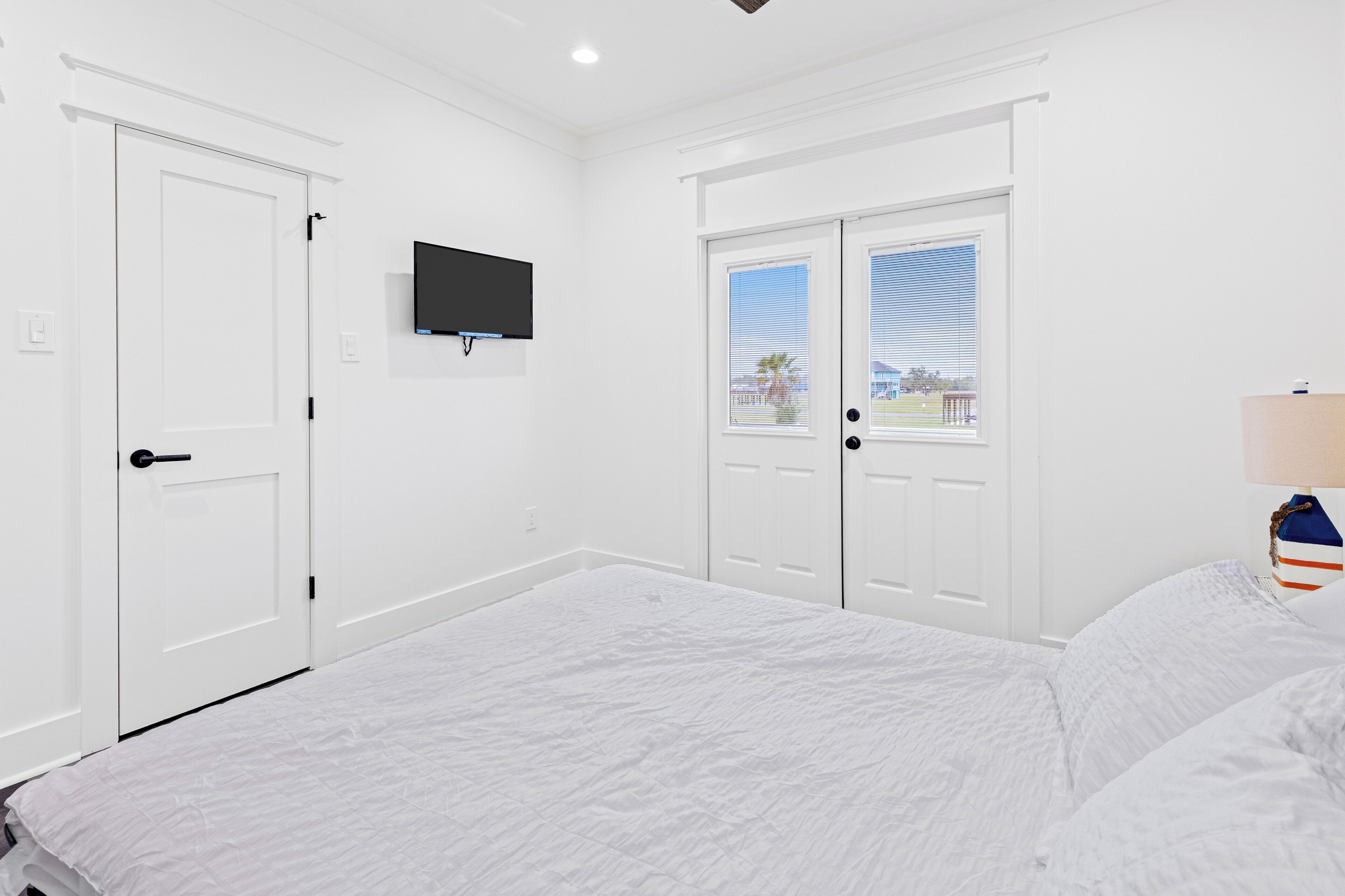 A bedroom with a large bed, white walls, a flat-screen TV mounted above a door, and a window with blinds.
