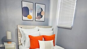 Un dormitorio con cama, almohadas, una mesita de noche, lámpara, ventana con persianas y arte abstracto en la pared.