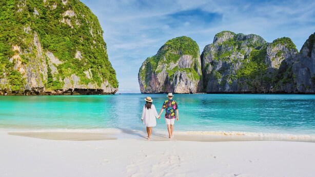 Un couple se promène sur une plage de sable blanc, avec une eau turquoise et limpide, et des falaises verdoyantes en arrière-plan.
