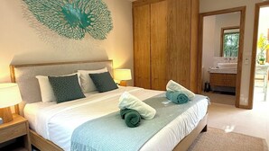 A bedroom with a bed, nightstands, a wooden wardrobe, a bathroom with a sink, and a decorative wall piece. - Luxury 3BR Pool Villa near Blue Tree - Bang Tao (Si Sunthon)