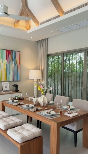 A modern dining room with a wooden table, cushioned benches, and a large window with a view of bamboo trees. - Luxury 3BR Pool Villa near Blue Tree - Bang Tao (Si Sunthon)