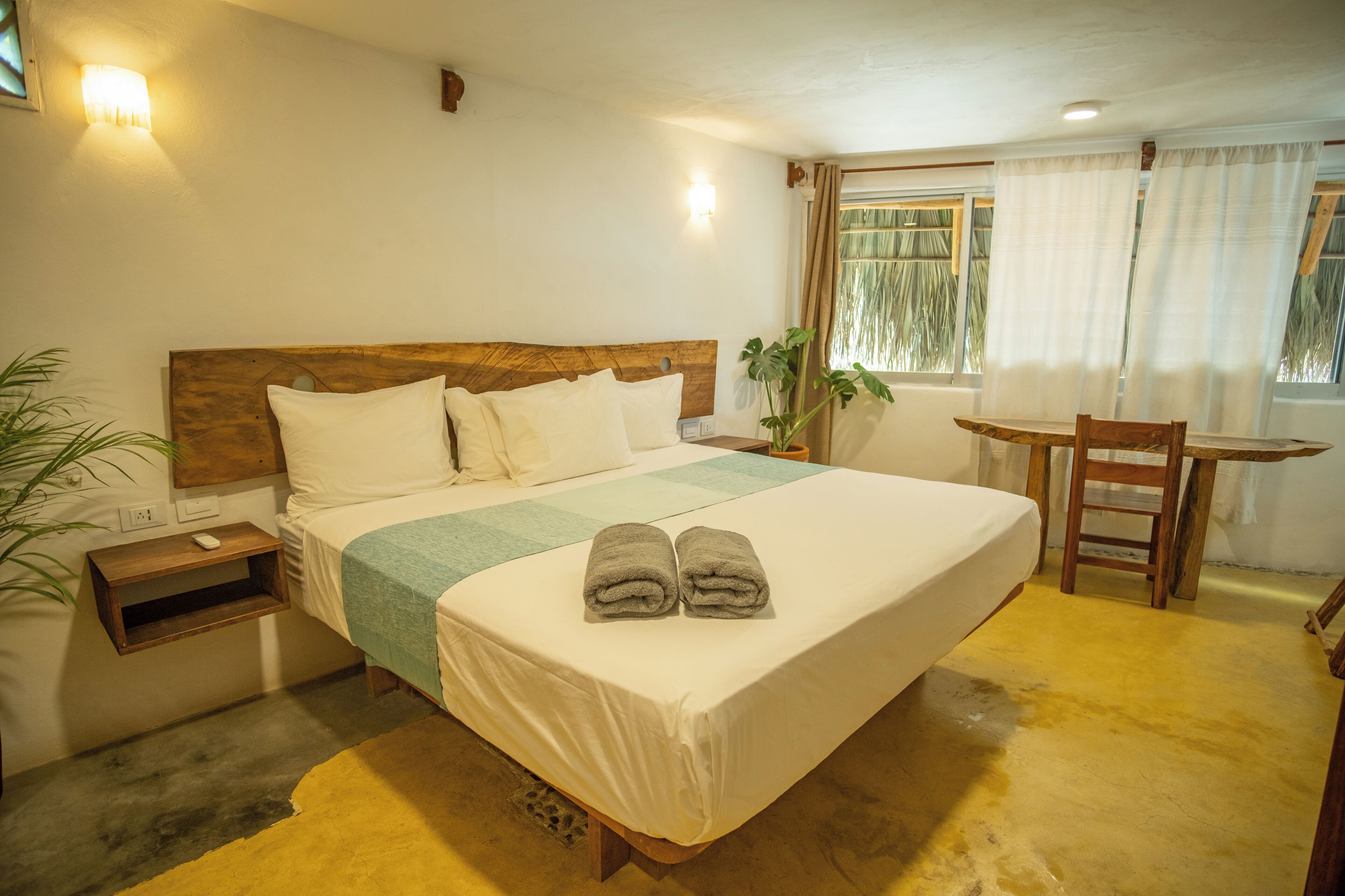 Une chambre d’hôtel avec un grand lit, une table de chevet, une chaise, une petite table et une fenêtre avec des rideaux.