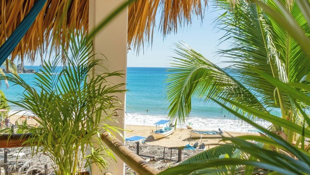Blick auf einen Strand vom Balkon mit einem Reetdach, einem Blumentopf und Palmenblättern.