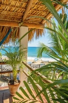 Vista a la playa desde un balcón con techo de paja, una planta en maceta y hojas de palmera.