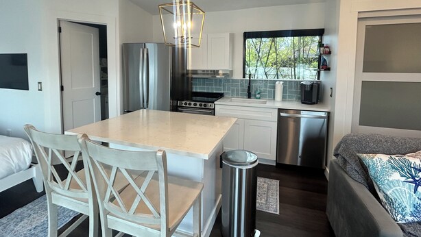 A compact kitchen with a central island, stainless steel appliances, and a window providing a view of greenery. - Tiny Mansion in Naples. 5 mins to Beach (Naples)