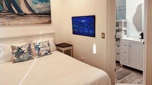 A bedroom with a bed, a nightstand, a painting of a cityscape, a TV, and a bathroom with a mirror and shelves. - Tiny Mansion in Naples. 5 mins to Beach (Naples)