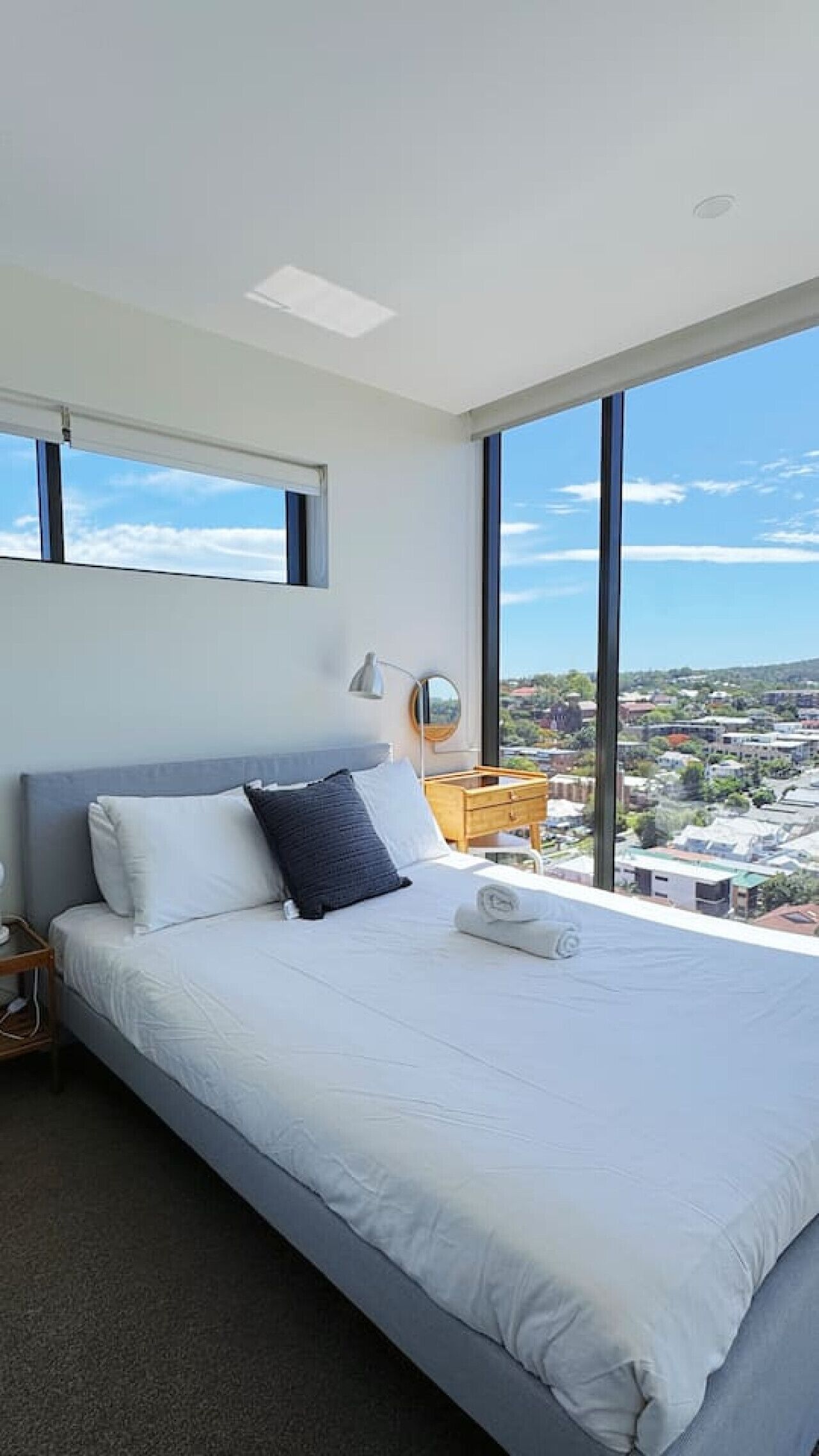 A modern bedroom with a large bed, a bedside table, a mirror, and a view of the city through large windows.