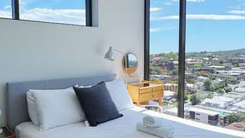 A modern bedroom with a large bed, a bedside table, a mirror, and a view of the city through large windows.