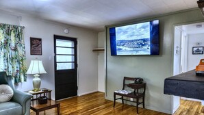 A living room with a wooden floor, a sofa, a chair, a lamp, a television mounted on the wall, and a door. - Cozy 2-bedroom home in Oak Ridge - 1940s themed (Oak Ridge)