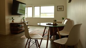 Ein Wohnzimmer mit Fernseher, einem runden Glastisch, zwei weißen Stühlen, einer Couch und einem Fenster mit Tageslicht.