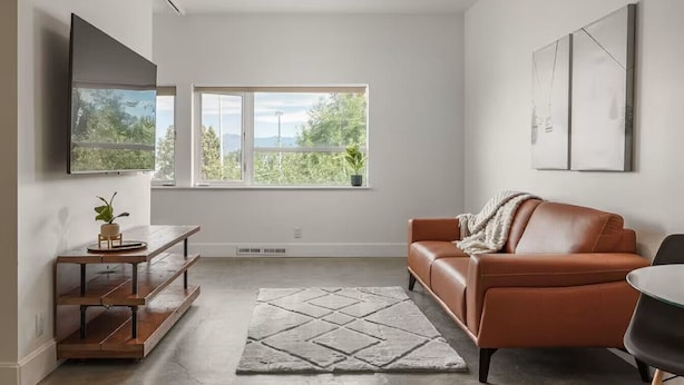 Ein modernes Wohnzimmer mit einem braunen Ledersofa, einem Holztisch, einem großen Flachbildfernseher an der Wand und einem großen Fenster mit Blick auf Grünflächen und Berge.