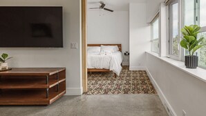 Ein modernes Schlafzimmer mit Bett, Deckenventilator, an der Wand montiertem Fernseher und einem Fenster mit Pflanze im Fensterbrett.