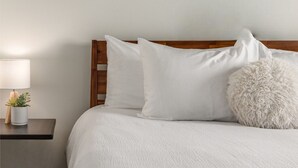 Ein ordentlich bezogenes Bett mit weißer Bettwäsche und Kissen, ein Holz-Kopfbrett und ein Nachttisch mit Lampe und einer kleinen Pflanze.