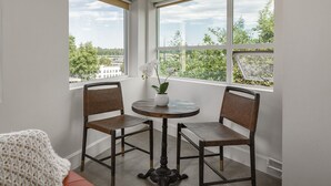 Habitación de esquina con dos sillas y una mesa, ventanas amplias con vistas a zonas verdes y edificios, y un objeto tejido blanco en el suelo.