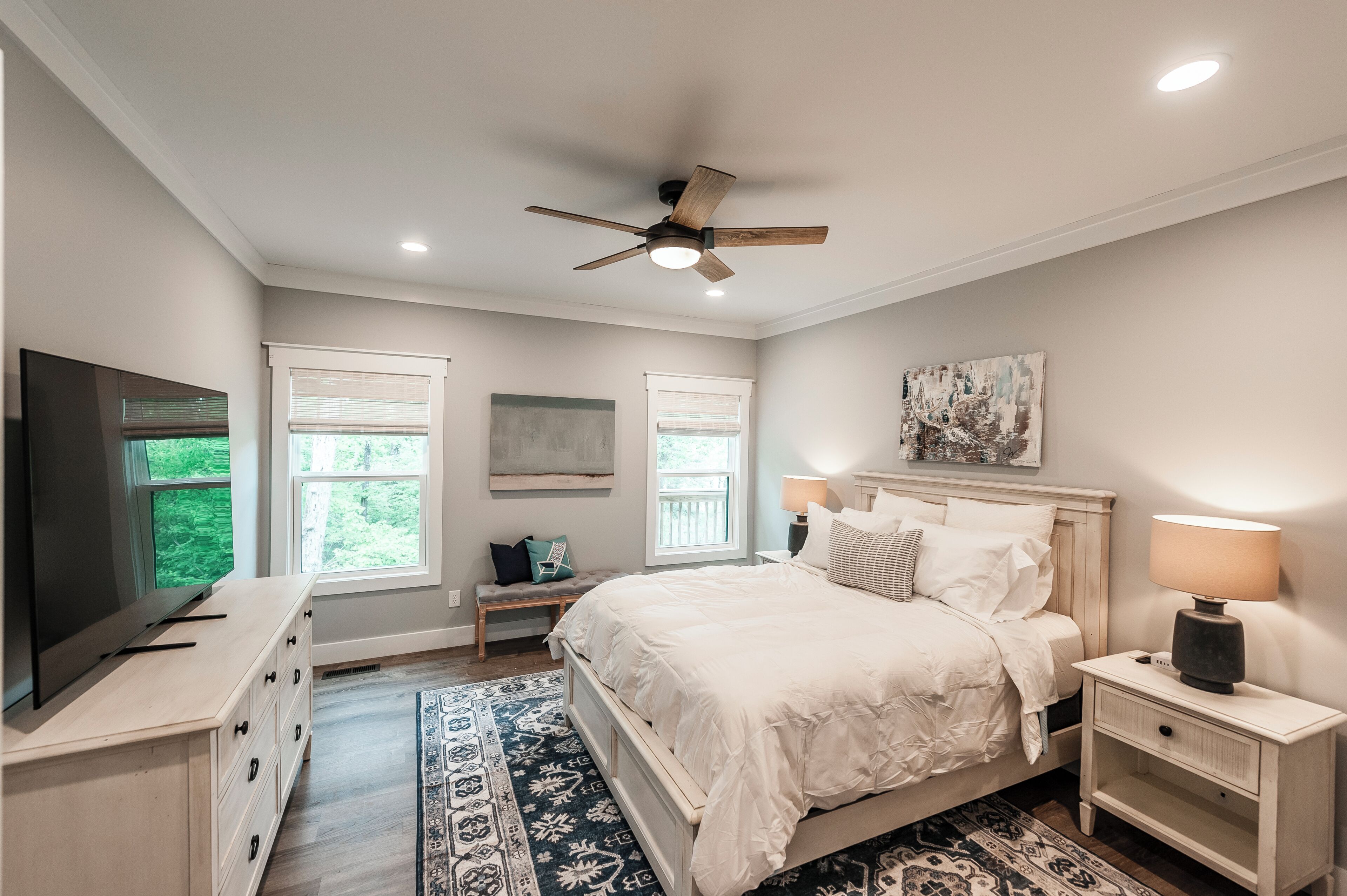 A bedroom with a bed, bedside tables, a ceiling fan, a TV, and windows.