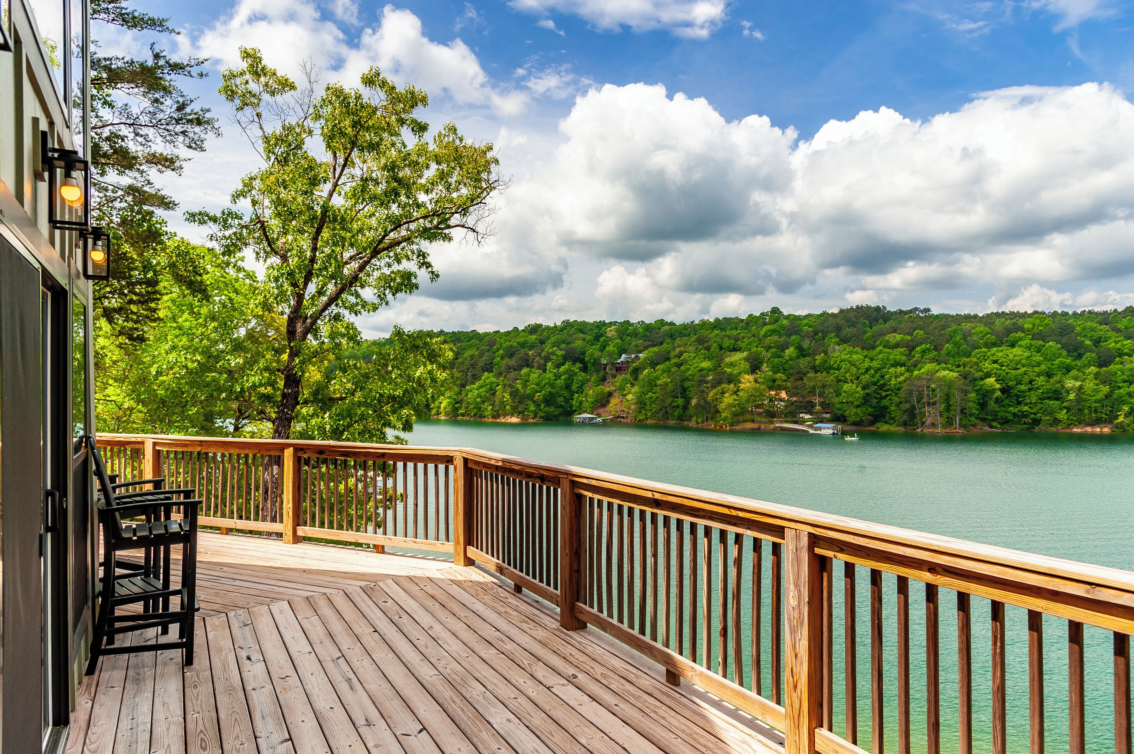 Una terraza de madera con barandilla que da a un lago, rodeada de árboles y un cielo despejado.