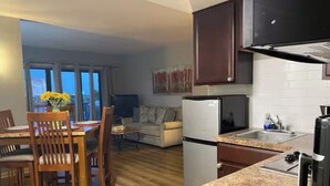Een moderne keuken met granieten aanrecht, een ingebouwde oven en een roestvrijstalen spoelbak. Er is een eethoek met houten stoelen en een tafel, en een woonkamer met een bank en salontafel.