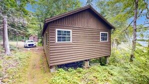 A wooden cabin with two windows, surrounded by trees and a path leading to a parked car. - Bay-View Getaway w/ Deck Between Camden & Belfast! (Northport)