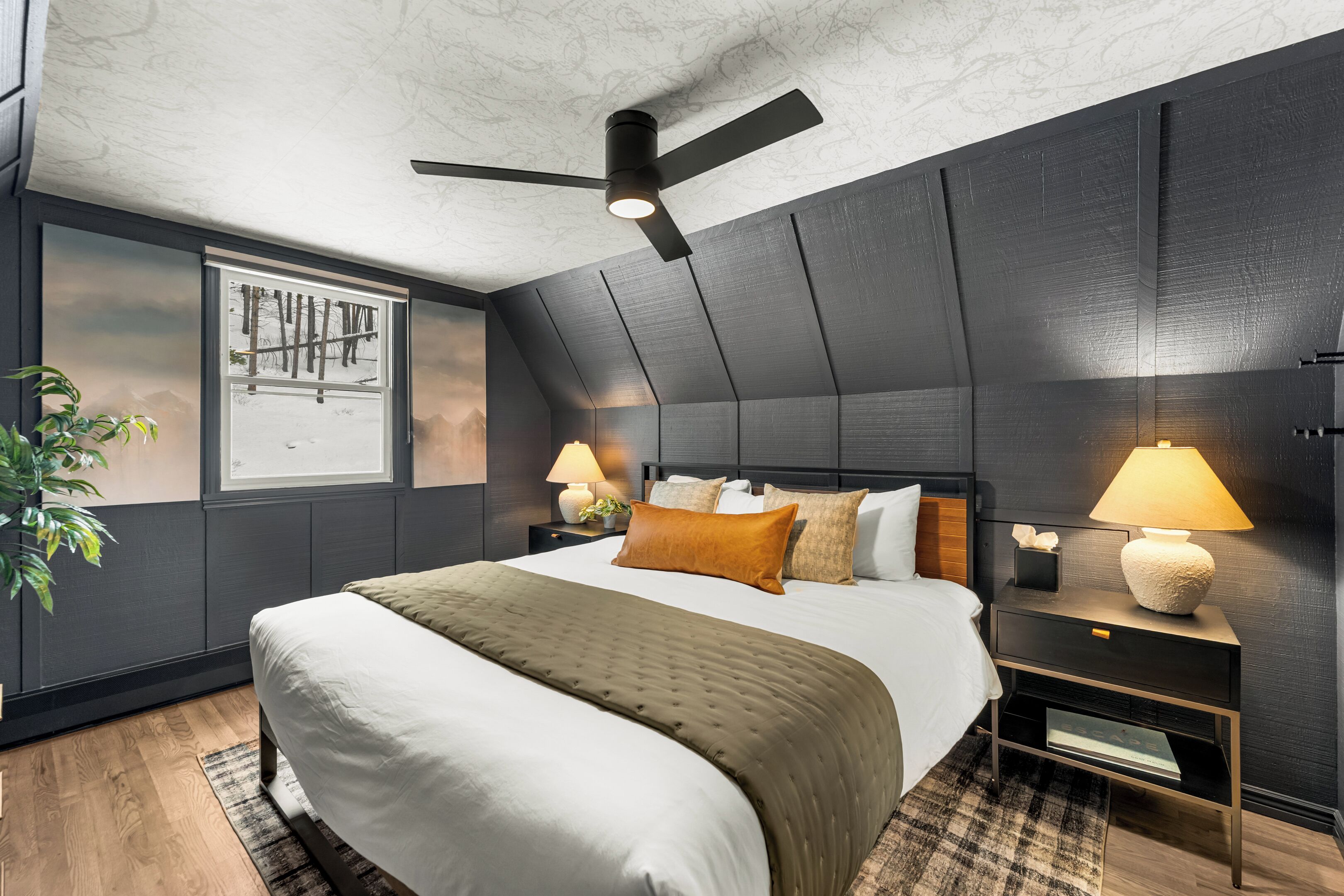 A modern bedroom with a large bed, dark walls, a ceiling fan, and two bedside tables with lamps.