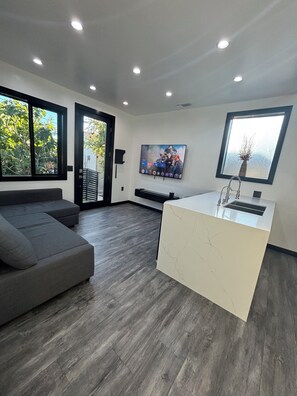 A modern living room with a grey sectional sofa, a white marble kitchen island, and a large flat-screen TV mounted on the wall. - Burbank - Starlight Hills Luxury Guest House (Burbank)