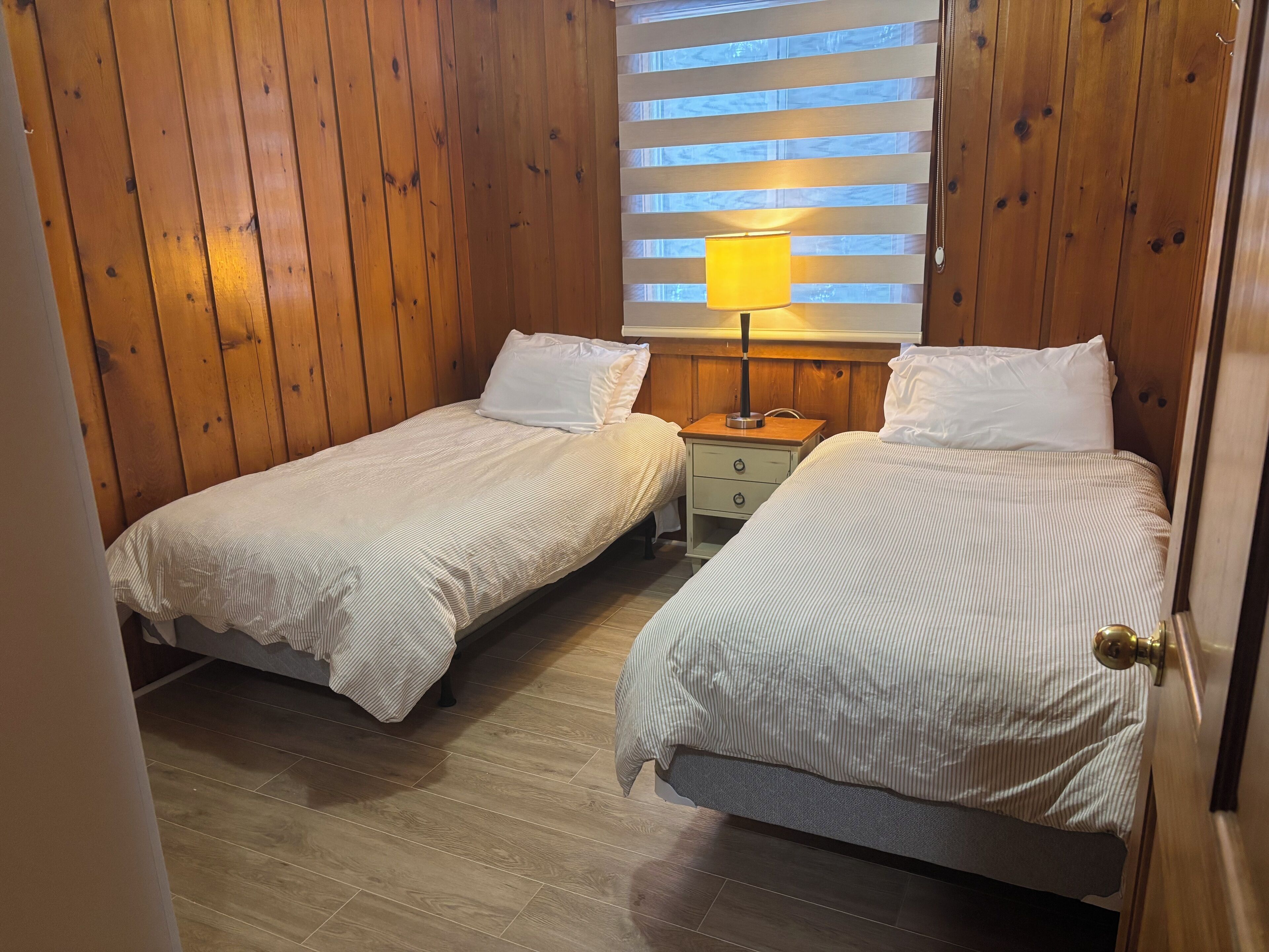 Una habitación con dos camas individuales, paredes de madera, una mesita de noche con lámpara y una ventana con persianas.