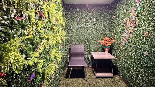 Une pièce avec un mur vert recouvert de feuillage, une petite table avec un vase et une chaise.