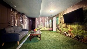 Une pièce comprenant un canapé, une table basse, un revêtement de sol en gazon synthétique, un téléviseur fixé au mur et une fresque représentant une forêt.