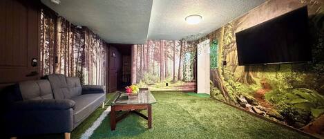 A room with a sofa, a coffee table, artificial grass flooring, a wall-mounted TV, and a forest mural.