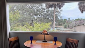 A round wooden table with two chairs, each with a leaf-patterned cushion, positioned by a window overlooking a natural landscape with trees and a building in the distance.