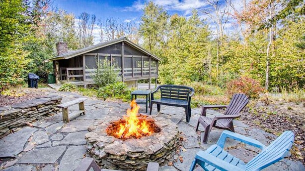 A stone fire pit surrounded by outdoor furniture, a wooden cabin, and a forested backdrop.