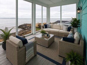 Una habitación iluminada por el sol con vistas al mar, que presenta muebles de mimbre, una mesa de centro de cristal y una planta en maceta.