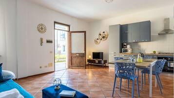 Ein modernes Wohnzimmer mit einer Couch, einem Couchtisch und einem Fernseher. Es gibt einen Essbereich mit Tisch und Stühlen. Der Raum verfügt über ein großes Fenster und eine Tür, die nach draußen führt.