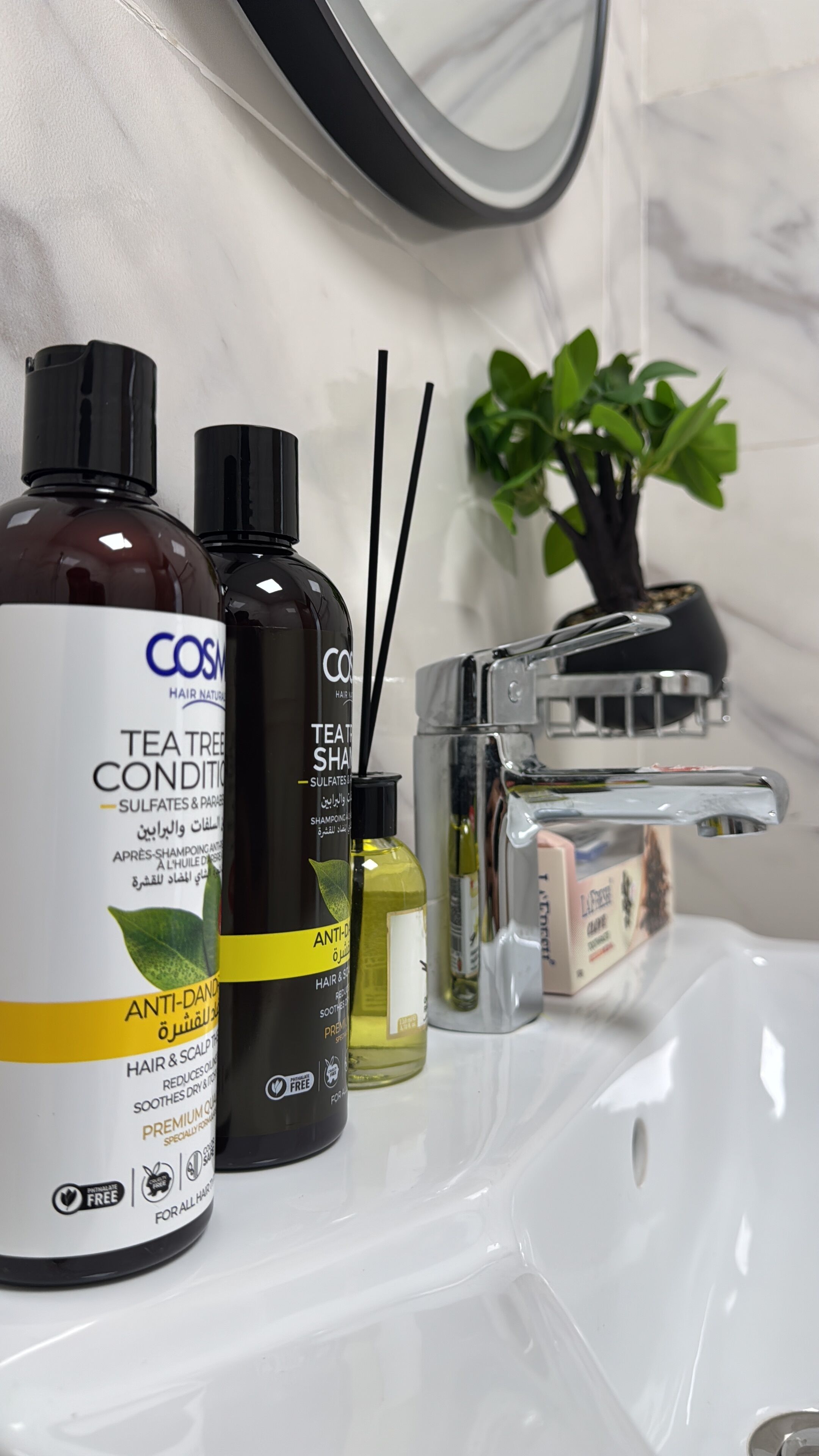Casa de banho com uma pia branca, um espelho e vários artigos de higiene pessoal, incluindo um frasco de condicionador de melaleuca, um frasco de champô e um frasco de condicionador.